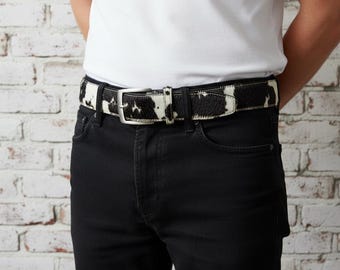 May include: A black and white cowhide belt with a silver buckle is worn over black jeans. The belt has a natural pattern of black spots on a white background. The person is wearing a white t-shirt.