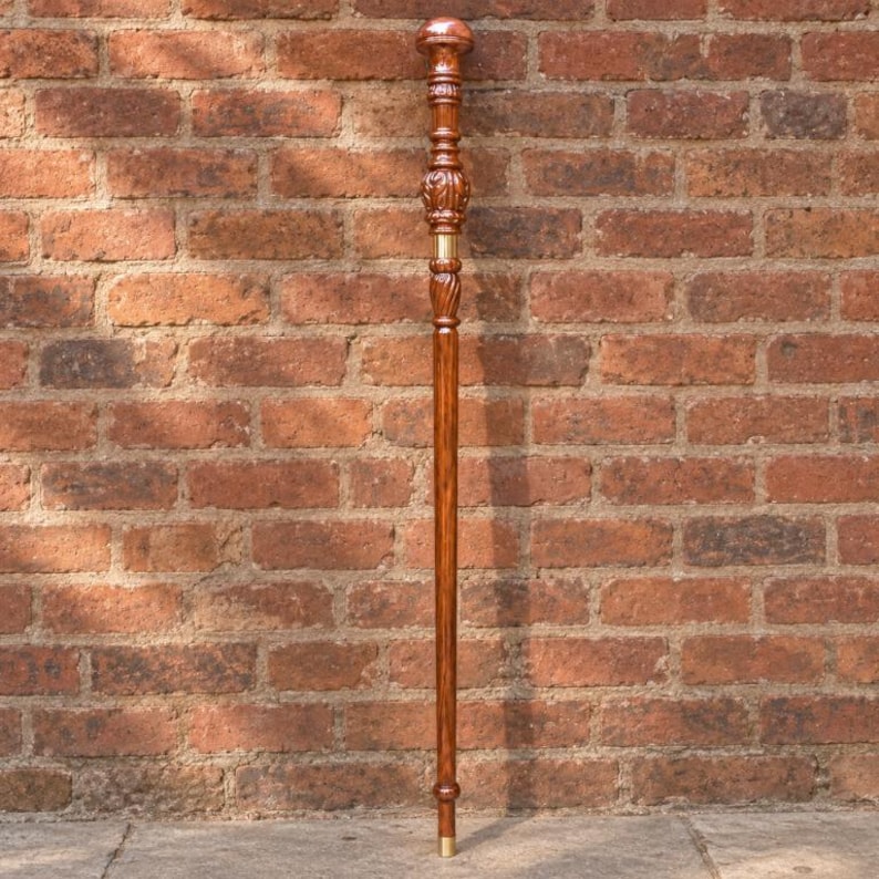 May include: A tall, slender walking stick crafted from polished brown wood with a visible grain. The handle features a rounded, carved top section with intricate detailing and a smooth, cylindrical shaft accented by a metallic gold-colored band.