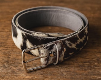 May include: A rolled belt with a cowhide pattern in black and white. The belt has a silver-colored buckle and a gray interior. The belt is made of leather and is suitable for everyday wear.