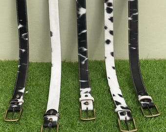 May include: Five leather belts with brass buckles. The belts have a cowhide pattern in black and white. One belt is solid white. The belts are arranged on a green surface.