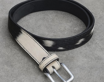 May include: A black and white cowhide belt with a silver-colored buckle. The belt features a black leather backing and a cream-colored strap with black and white spotted markings. The buckle is a standard square design.