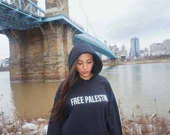 May include: A person wearing a black hooded sweatshirt with the text "FREE PALESTINE" in white lettering. The person is standing in front of a bridge over a river.