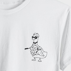 Puede incluir: Camiseta blanca con una ilustración en blanco y negro de un ganso con gafas de sol, un sombrero y un palo de golf. El ganso tiene un cuerpo estampado. La camiseta está hecha de un material suave y cómodo.