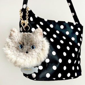 May include: A black shoulder bag with white polka dots, adorned with a fluffy grey cat-shaped charm with blue eyes. A gold chain and a tag with the name "Remy" are attached.