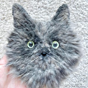 May include: A handmade plush cat face in shades of grey and brown, with green eyes and white whiskers. The cat has a fluffy texture and pointed ears. The cat's nose is small and black. The item is a handcrafted cat.