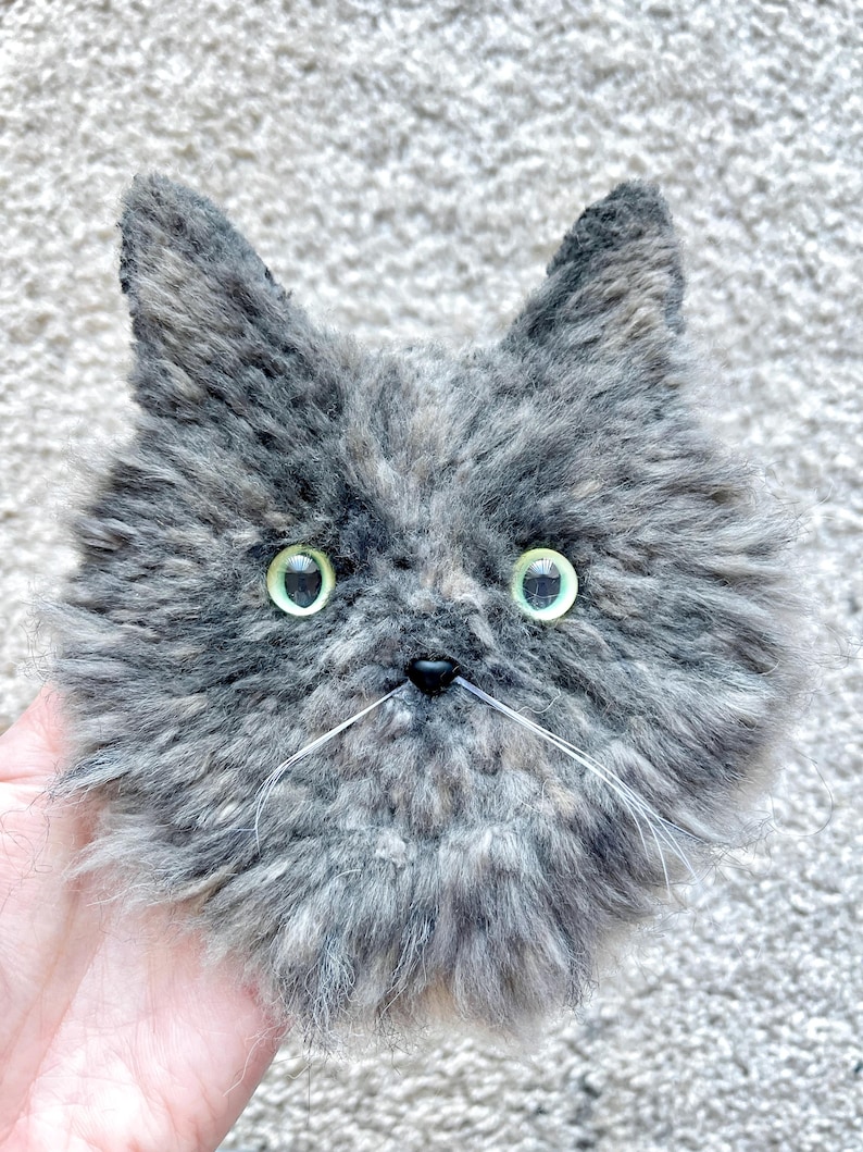 May include: A handmade plush cat face in shades of grey and brown, with green eyes and white whiskers. The cat has a fluffy texture and pointed ears. The cat's nose is small and black. The item is a handcrafted cat.