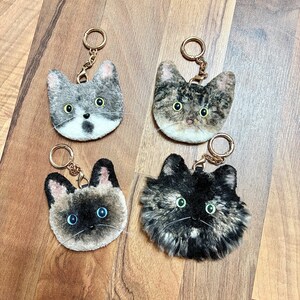 May include: Four cat-shaped keyrings with gold-coloured rings. The keyrings feature different cat fur patterns: grey tabby, brown tabby, Siamese, and tortoiseshell. Each keyring has a detailed cat face with eyes and a nose.