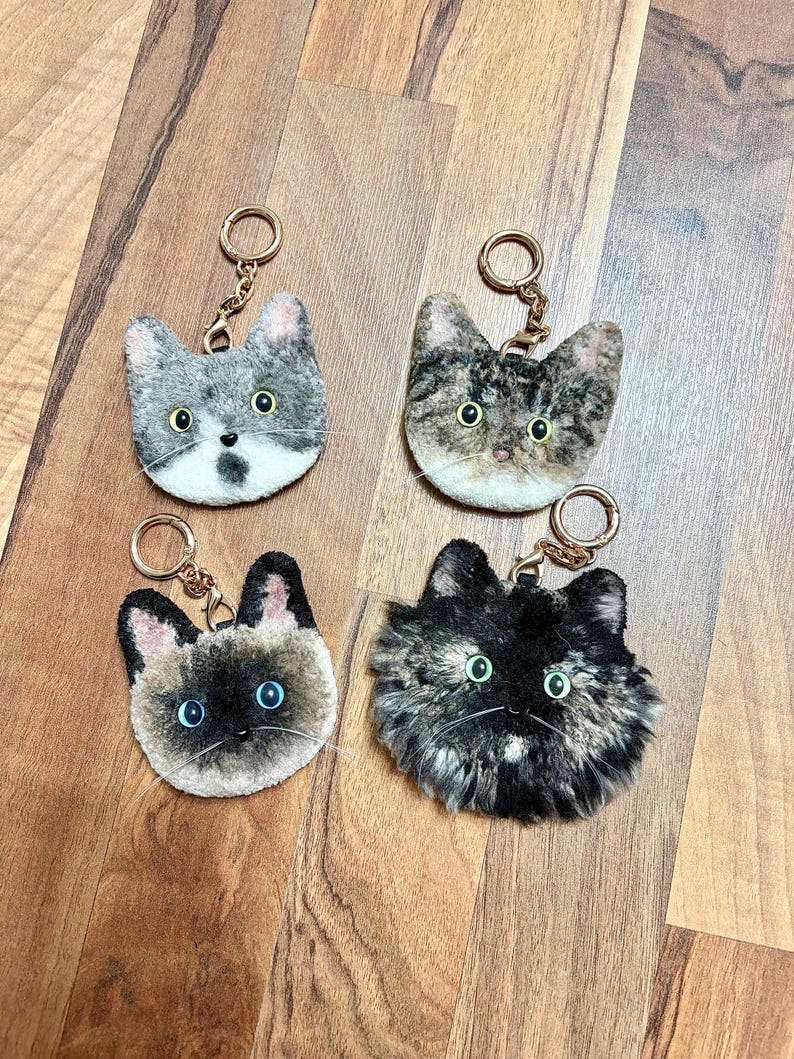 May include: Four cat-shaped keyrings with gold-coloured rings. The keyrings feature different cat fur patterns: grey tabby, brown tabby, Siamese, and tortoiseshell. Each keyring has a detailed cat face with eyes and a nose.