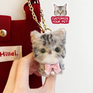 May include: A small, gray and white cat plush toy with a pink bow and a pearl charm. The plush toy is hanging from a gold chain and is attached to a red bag with the word "Hilldi." printed on it. The image also includes a sticker with a cat face and the text "Customize Your Pet!"