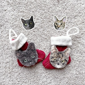 Puede incluir: Dos calcetines de Navidad rojos y blancos con caras de gato. Los calcetines están colgados en una superficie blanca y esponjosa. Las caras de los gatos son grises y marrones.