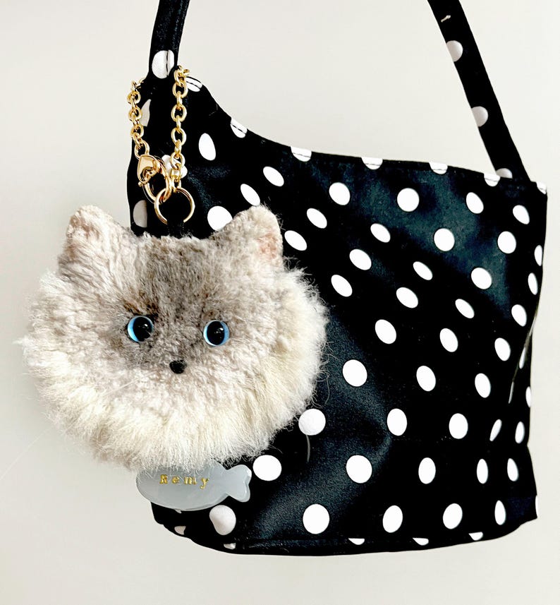 May include: A black shoulder bag with white polka dots, adorned with a fluffy grey cat-shaped charm with blue eyes. A gold chain and a tag with the name "Remy" are attached.