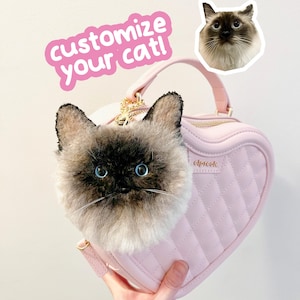 May include: A pink heart-shaped handbag with a plush cat face attached. The bag has a quilted pattern and a gold zip. The text "customize your cat!" is in a pink sticker. A cat sticker is also visible.