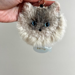 May include: A fluffy cat keyring with a gray and white face and large blue eyes. The keyring has a gold-coloured ring and a small tag that reads "Remy". The cat's face is detailed with a black nose and whiskers.