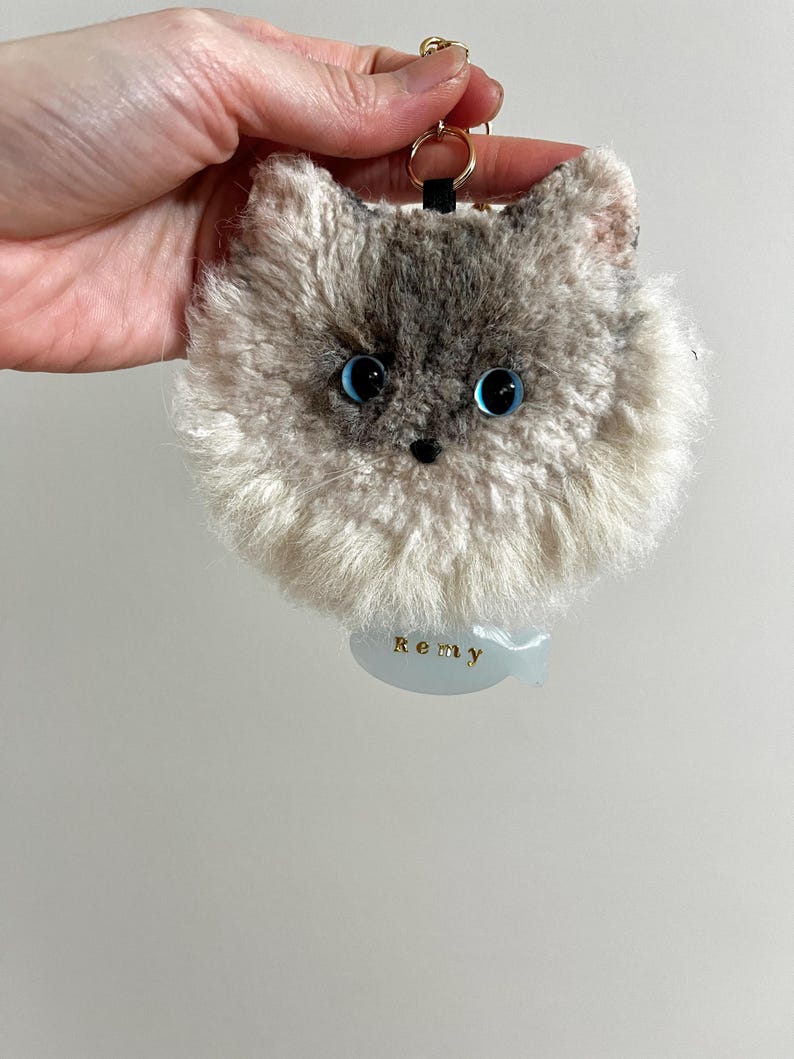 May include: A fluffy cat keyring with a gray and white face and large blue eyes. The keyring has a gold-coloured ring and a small tag that reads "Remy". The cat's face is detailed with a black nose and whiskers.