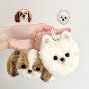 May include: Two plush dog ornaments, one a Shih Tzu with brown and white fur, the other a white Pomeranian. Both have black eyes and small pink noses. The ornaments are held by a hand, with two dog face stickers in the background.