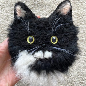 May include: A fluffy cat face made of black and white material. The cat has yellow eyes, long white whiskers, and pink inner ears. The cat's face is round and fluffy, with a friendly expression. The cat's muzzle and chest are white.