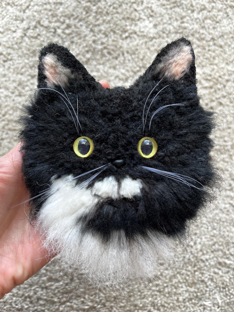 May include: A fluffy cat face made of black and white material. The cat has yellow eyes, long white whiskers, and pink inner ears. The cat's face is round and fluffy, with a friendly expression. The cat's muzzle and chest are white.