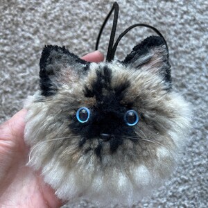 May include: A fluffy cat head ornament with bright blue eyes, a black nose, and whiskers. It has a black cord for hanging. The fur is a mix of black, brown, and cream.