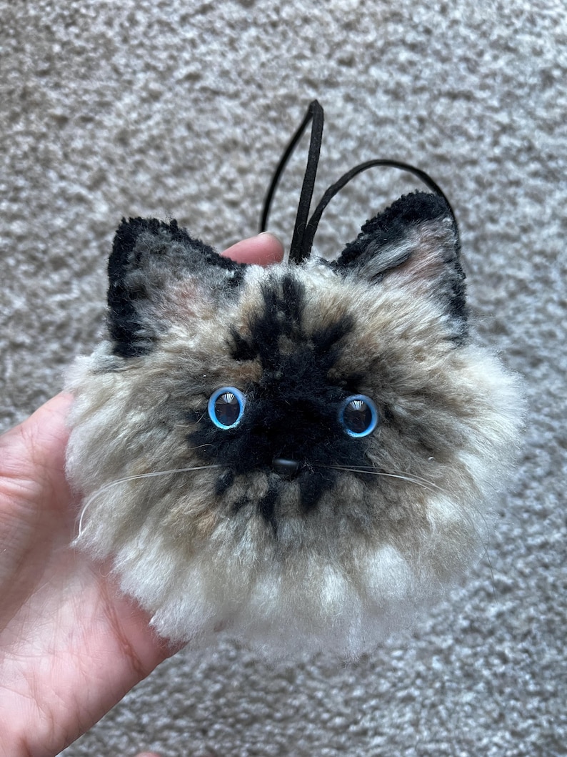 May include: A fluffy cat head ornament with bright blue eyes, a black nose, and whiskers. It has a black cord for hanging. The fur is a mix of black, brown, and cream.