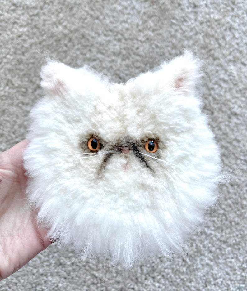 May include: A fluffy white cat face with orange eyes and a grumpy expression. The cat has a dark grey nose and a small, dark grey line above the eyes. The cat's fur is long and shaggy, giving it a soft appearance.