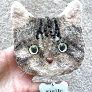 May include: A soft toy cat's face with green eyes, a pink nose, and white whiskers. The cat has a grey and brown tabby pattern. A pale blue bone-shaped tag with the name "winlie" is attached below.