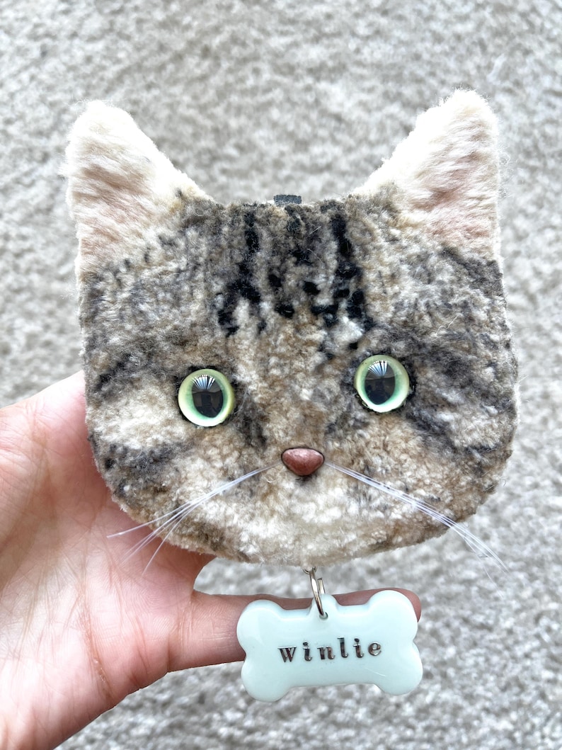May include: A soft toy cat's face with green eyes, a pink nose, and white whiskers. The cat has a grey and brown tabby pattern. A pale blue bone-shaped tag with the name "winlie" is attached below.