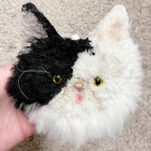 May include: A handmade plush cat head with black and white fur. The cat has yellow eyes, a pink nose, and a fluffy white muzzle. The black fur covers one ear and half of the head, while the other half is white.