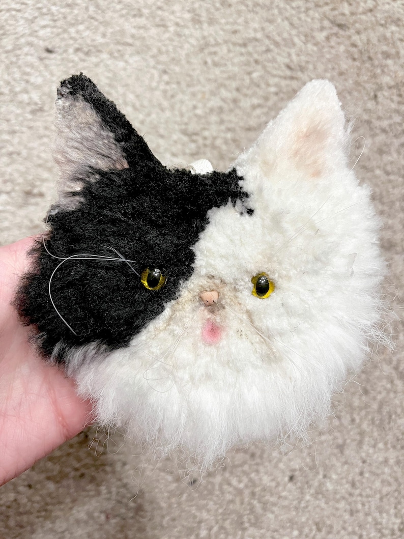 May include: A handmade plush cat head with black and white fur. The cat has yellow eyes, a pink nose, and a fluffy white muzzle. The black fur covers one ear and half of the head, while the other half is white.