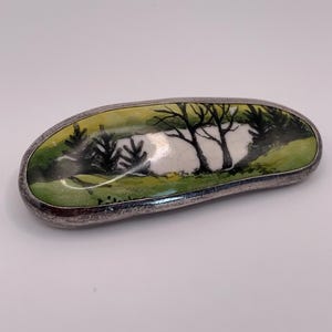 Pode incluir: Broche oval com uma cena de paisagem pintada. O broche apresenta uma moldura de metal prateado em torno de uma cena pintada de árvores e água. A paisagem é pintada em tons de verde, branco e preto.