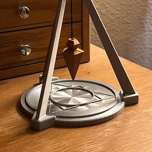 May include: A gray 3D printed pendulum with a wooden bob hanging from a black string. The pendulum is suspended from a triangular frame and sits on a circular base with a geometric design.