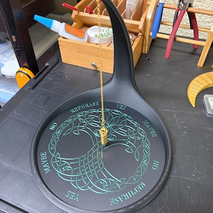 A black Tree of Life pendulum stand holds a gold chain and fob, set on a cluttered workbench with tools in the background.