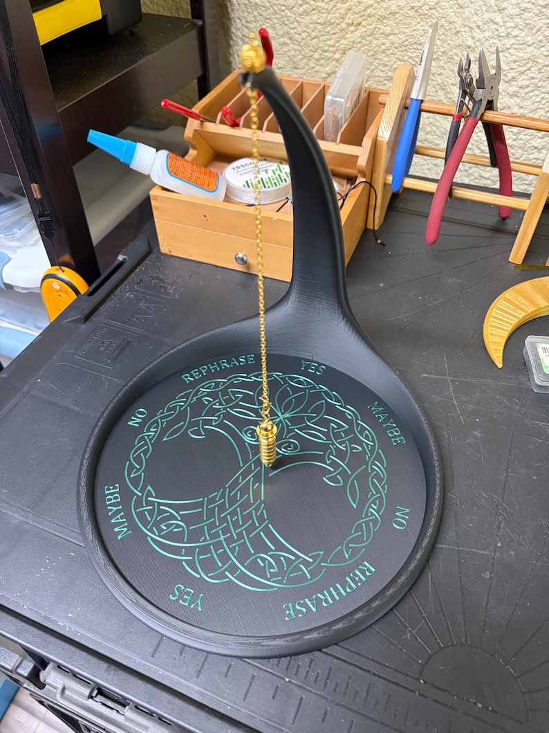 A black Tree of Life pendulum stand holds a gold chain and fob, set on a cluttered workbench with tools in the background.