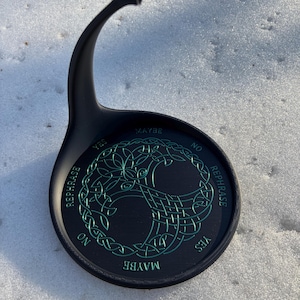 A black pendulum stand with green Tree of Life artwork and divination text is placed outdoors in the snow.