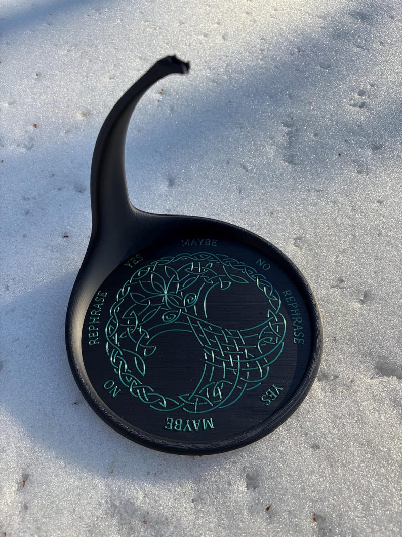 A black pendulum stand with green Tree of Life artwork and divination text is placed outdoors in the snow.