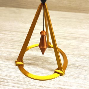 May include: A gold-colored metal pyramid with a round base and a black ball at the top. A copper-colored pendulum hangs from the top of the pyramid.