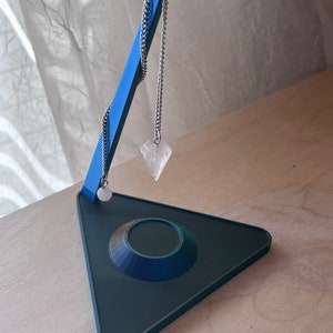May include: A blue and green 3D printed pendulum stand with a clear crystal pendulum hanging from a silver chain.