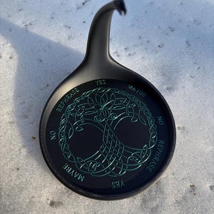 A black divination stand featuring a green Tree of Life pattern sits upright on a layer of fresh snow.