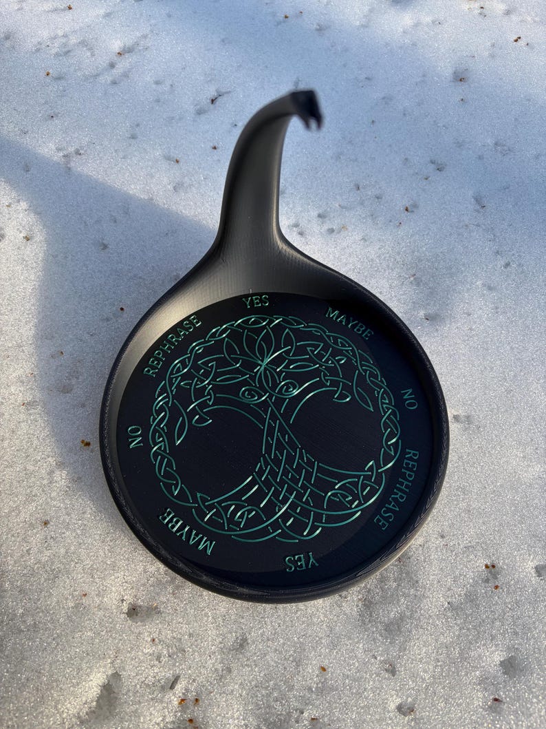 A black divination stand featuring a green Tree of Life pattern sits upright on a layer of fresh snow.