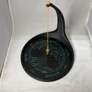 A gold chain with a metal pendulum hangs suspended over a black divination stand marked with a green Tree of Life motif.