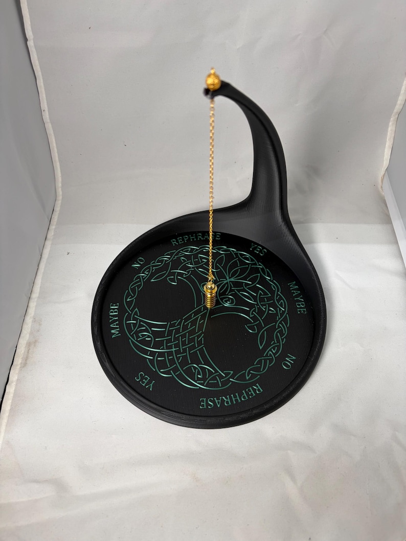A gold chain with a metal pendulum hangs suspended over a black divination stand marked with a green Tree of Life motif.