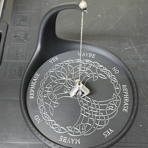 A black pendulum stand with white engraving displays a silver-colored pendulum dangling from its top arm over a Tree of Life design.