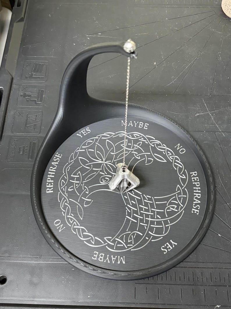 A black pendulum stand with white engraving displays a silver-colored pendulum dangling from its top arm over a Tree of Life design.