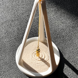 May include: A white zen garden set with a gold-colored pendulum hanging from a white frame. The sand is beige with circular patterns. The set is on a textured gray surface.