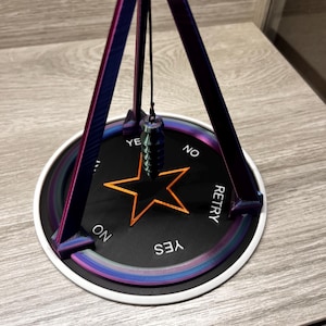 May include: A black and white pendulum with a star design and the words "YES", "NO", "RETRY" on a circular base. The base is made of a 3D printed material with a purple and blue iridescent finish.