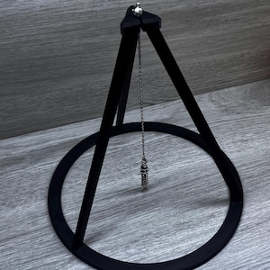 May include: A black pendulum stand with a circular base and three triangular supports. A silver chain hangs from the top, suspending a silver pendulum. The stand is made of a matte black material.