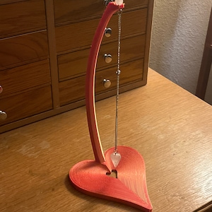 Puede incluir: Un soporte en forma de corazón rosa con un brazo de madera curvado que sostiene un péndulo de cuarzo transparente en una cadena plateada.