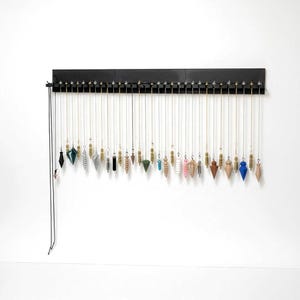 May include: A black horizontal wall-mounted display rack holding a collection of thirty-two dowsing pendulums. Each pendulum hangs from a thin metal chain, featuring various shapes, colors, and materials including wood, crystal, and metal.