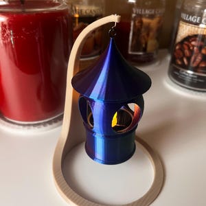 May include: A blue and gold candle lantern with a decorative design hangs from a wooden stand. The lantern has a round base and a pointed top. The stand is made of wood and has a circular base.