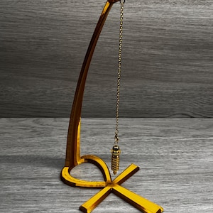 May include: A gold-coloured pendulum on a stand shaped like the Ankh symbol. The pendulum is hanging from a chain and is suspended above the Ankh.