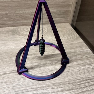 May include: A purple and green iridescent metal pendulum with a spiral design hangs from a metal stand. The stand is a triangular pyramid with a circular base.